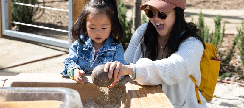 A parent scoops sand with a young child outdoors.