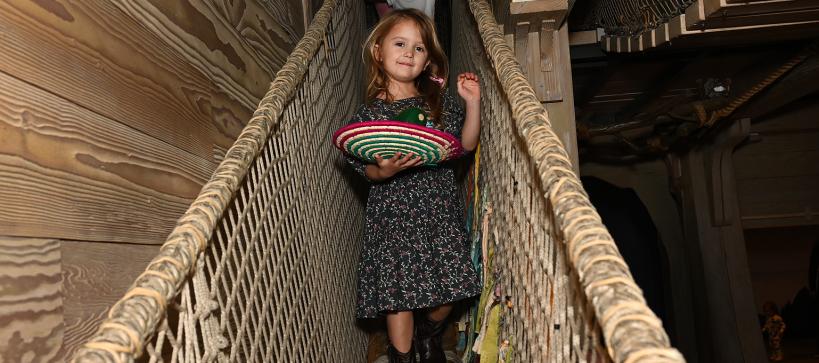 A child walks down a rope bridge holding a colorful basket.
