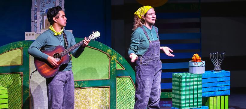 A man palys guitar and an actor on stage performing a Hanukkah story.