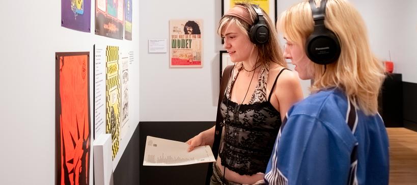 Two young girls look at comic posters with headphones on, in a gallery.