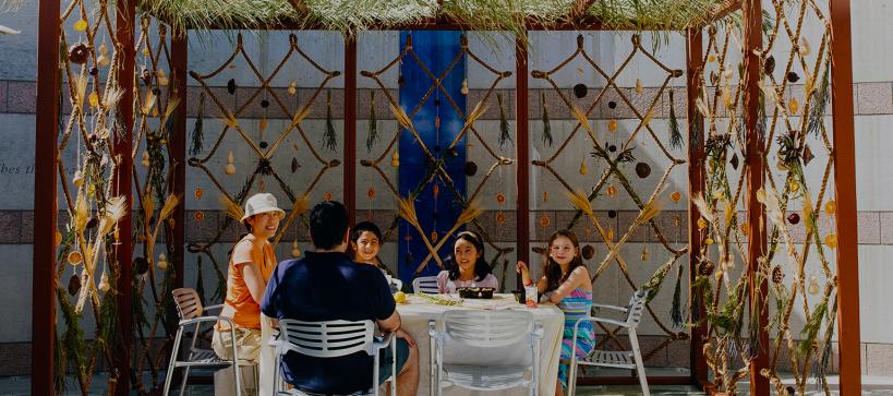 A family sits under a shelter made form palm leaves and hanging citrus fruit—a Sukkah