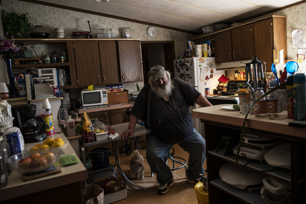 A light skinned adult man with a long grey beard is pictured in his kitchen sitting on a stool.