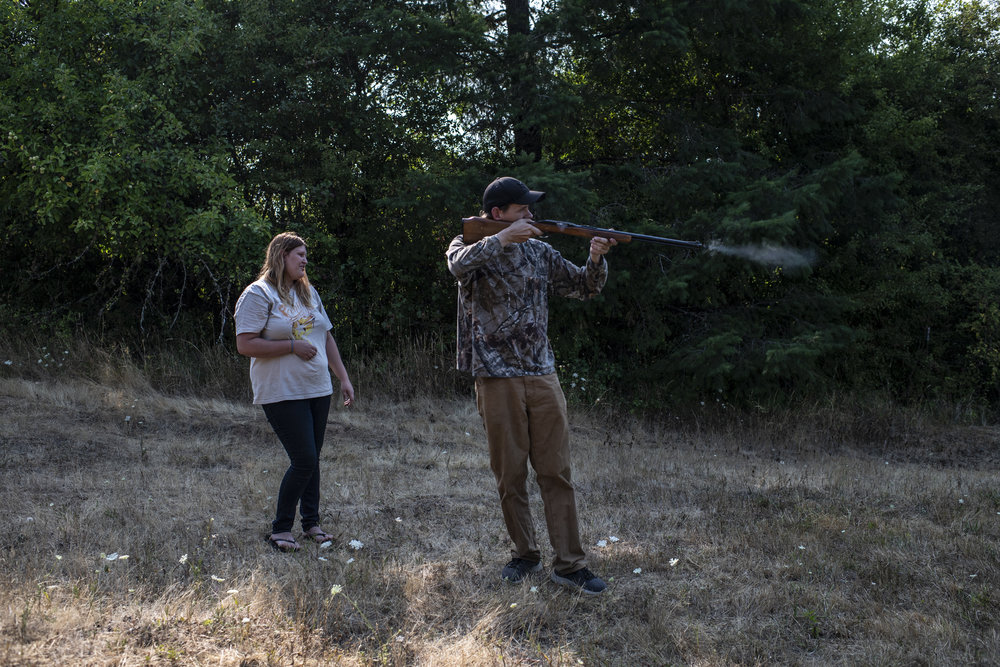 An adult man with light skin tone stands in a field with a gun practicing target shooting. Smoke rises from the gun’s barrel and a blond woman with light skin tone looks on smiling.