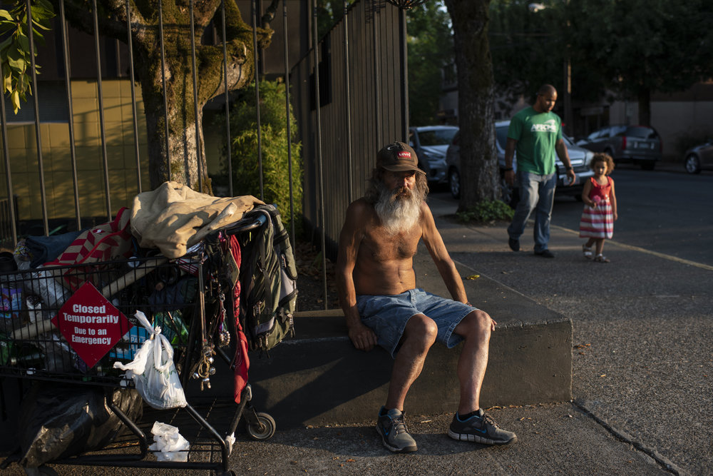 A shirtless adult man with light skin tone sits on the curb next to a shopping cart containing all of his belongings.