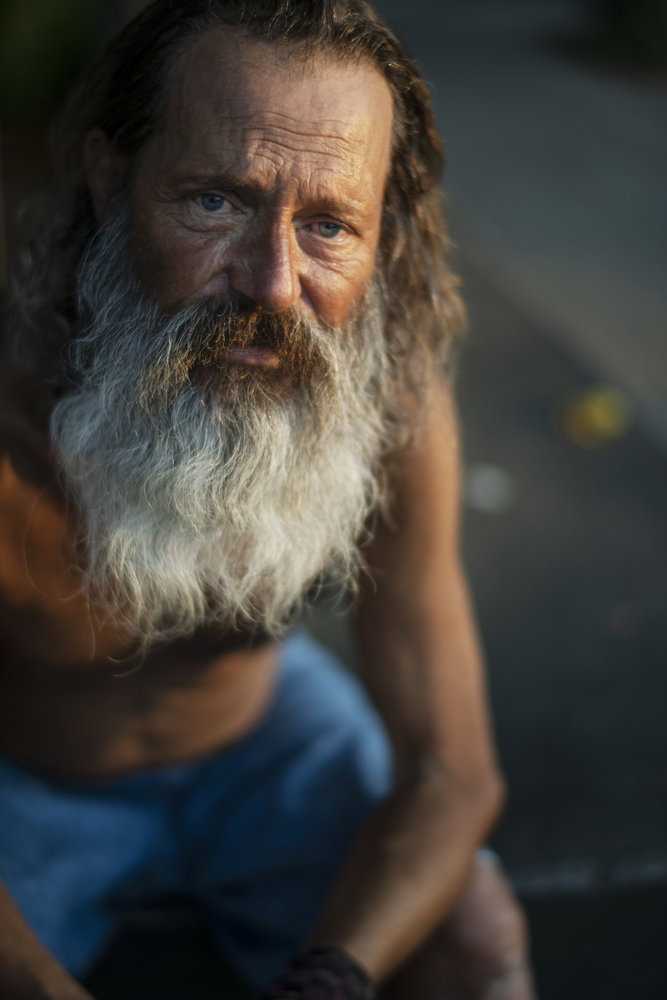 An adult man with light skin tone and a large grey beard sits shirtless looking solemnly at the camera.