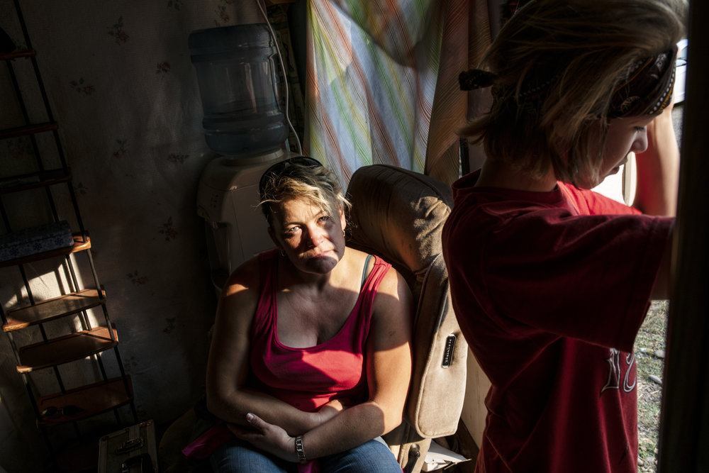 An adult woman with light skin tone is pictured inside a dark room sitting on a recliner. Her tween son with light skin tone peers out a window, pushing back a curtain, causing sunlight to break the darkness and splash across her face.