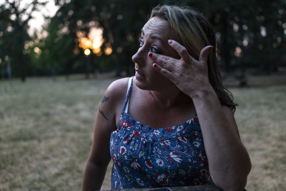 An adult woman with light skin tone is pictured at twilight sitting in a park wiping a tear from her eye.
