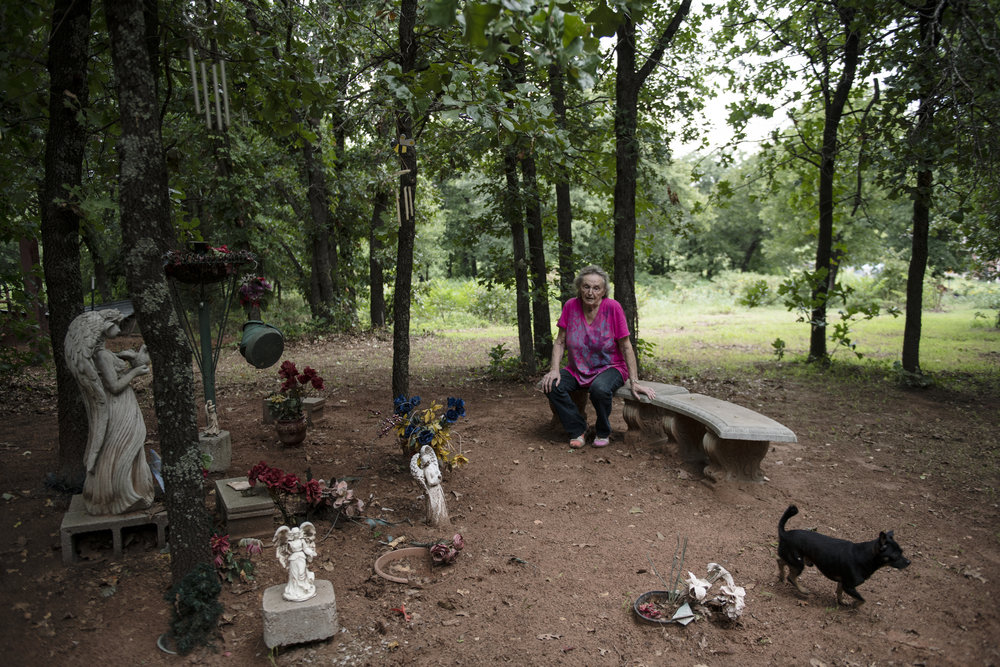 A light skinned older person sits outdoors on a stone bench surrounded by the graves of her dead children.