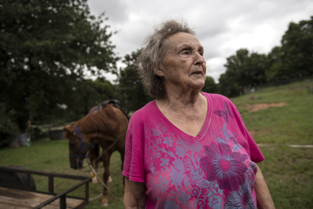 A light skinned older person wearing a pink shirt looks to the right of camera as though deep in thought. Behind her is a pasture where a brown mare grazes under an overcast sky.
