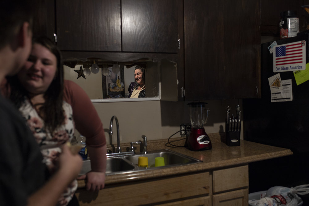 An adult with light skin tone stands in her kitchen wearing an apron smiling at her two teenage children, a boy and a girl with light skin tone.