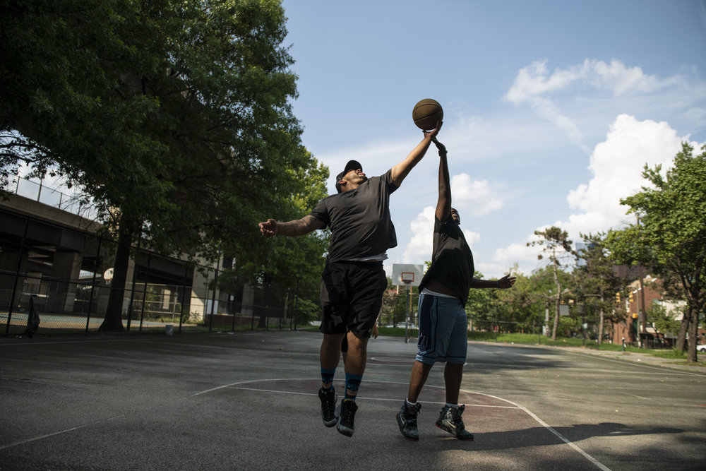Two adult men with medium and dark skin tones play basketball, their arms are outstretched in a motion that suggests they are literally and figuratively reaching for hope.