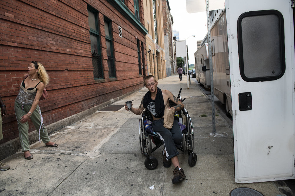 An adult man with light skin tone who uses a wheelchair is pictured on the sidewalk outside a safe needle exchange site. To his left stands an adult woman with medium skin tone and bright blond hair.