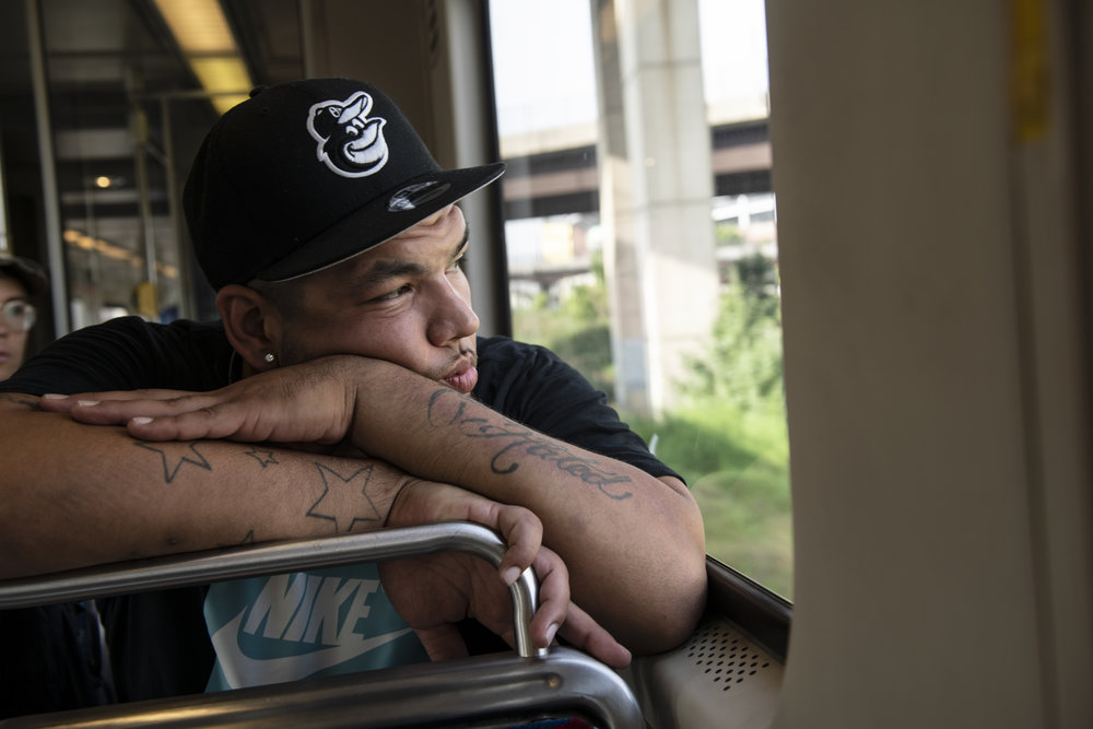 An adult man with medium skin tone is seated on public transit looking out of a window, his tattooed forearms are folded and resting on the back of a seat.
