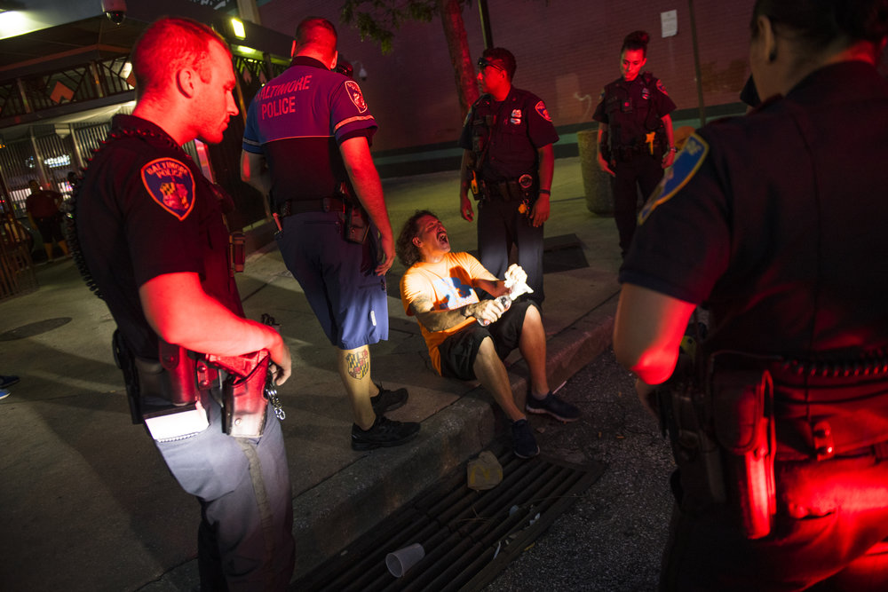 Police officers encircle an adult man with medium skin tone sitting on the pavement. The man is likely overdosing on drugs and his head is violently cocked back in anguish as the red glare of a police light jets across his face.