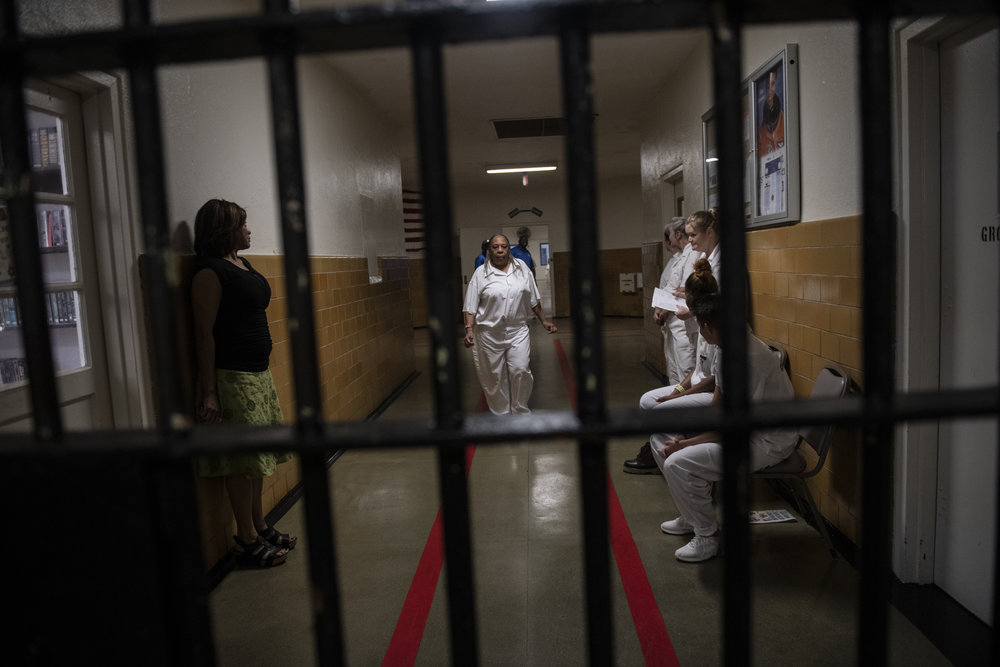 An older person with dark skin tone walks down the hallway of a women’s prison.