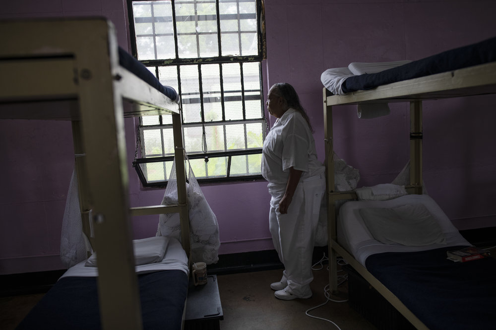 An older person with dark skin tone is pictured in a white jumpsuit looking out the window of a women’s prison dormitory.