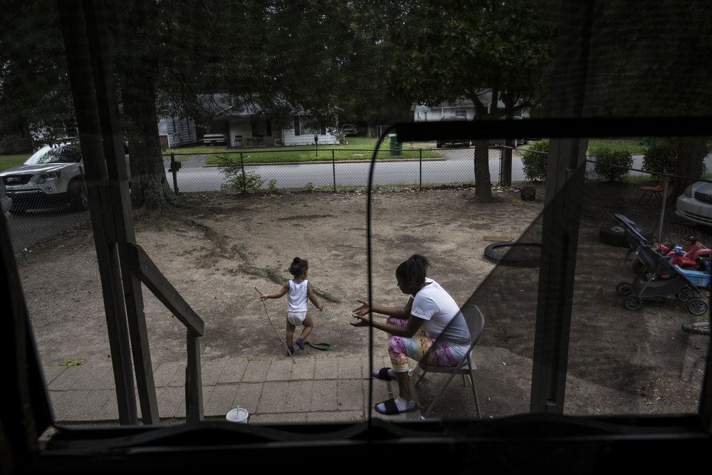 An adult woman with medium skin tone sits looking after her toddler in a front yard.
