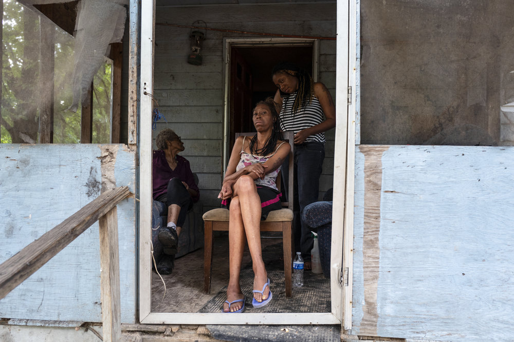 A multigenerational family of women with medium to dark skin tone sits on a porch together braiding hair.
