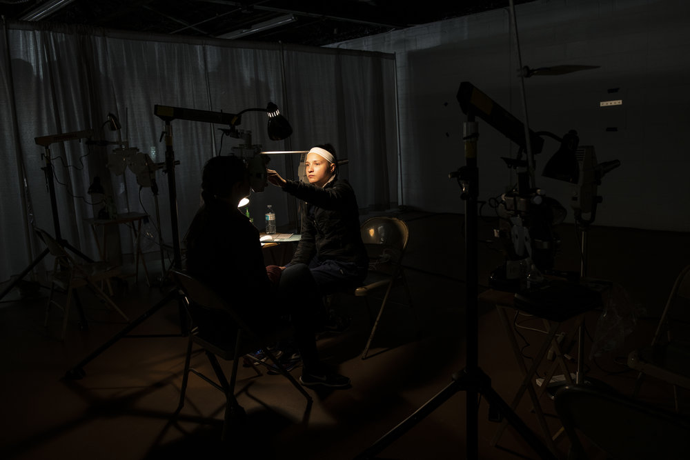 The room is very dark, obscuring the face of the woman receiving an eye exam. The darkness is broken by a light from a testing machine illuminating the face of the health care worker conducting the eye exam.