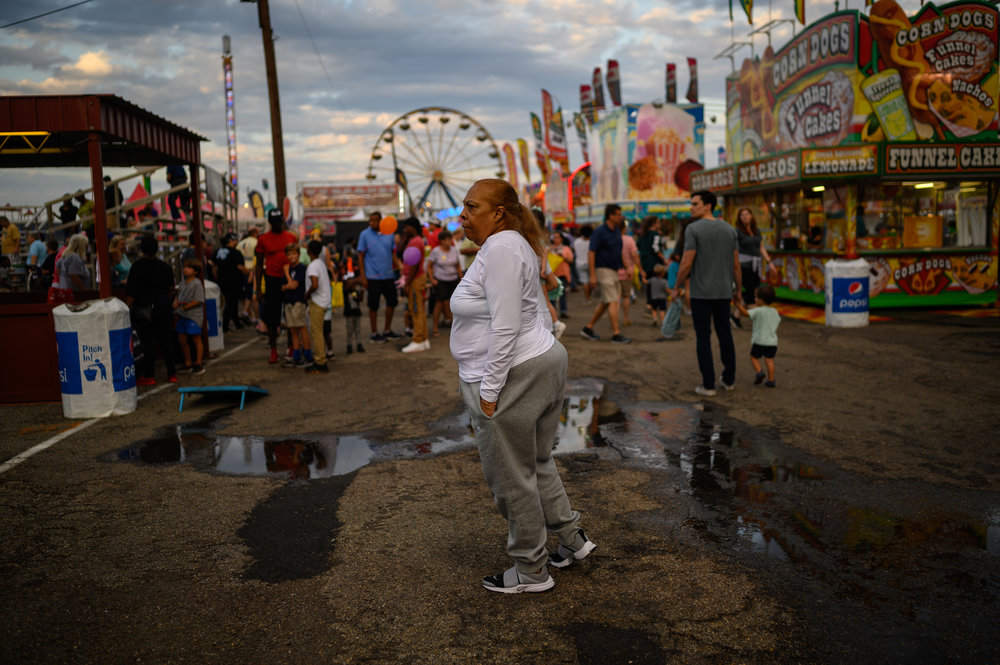 An older person with dark skin tone wearing causal clothing stands in the middle of a bustling fair ground.