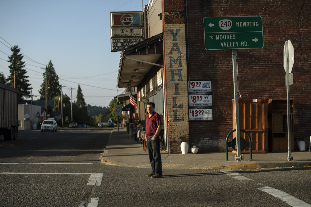 A light skinned adult man wearing a red t-shirt stands somberly in front of a brick general store.