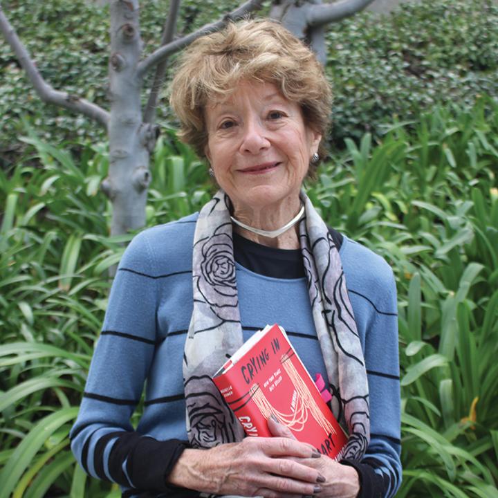 Photo of Carryl Carter sitting in front of lush, green plants. She is smiling at the camera and holding a red book.