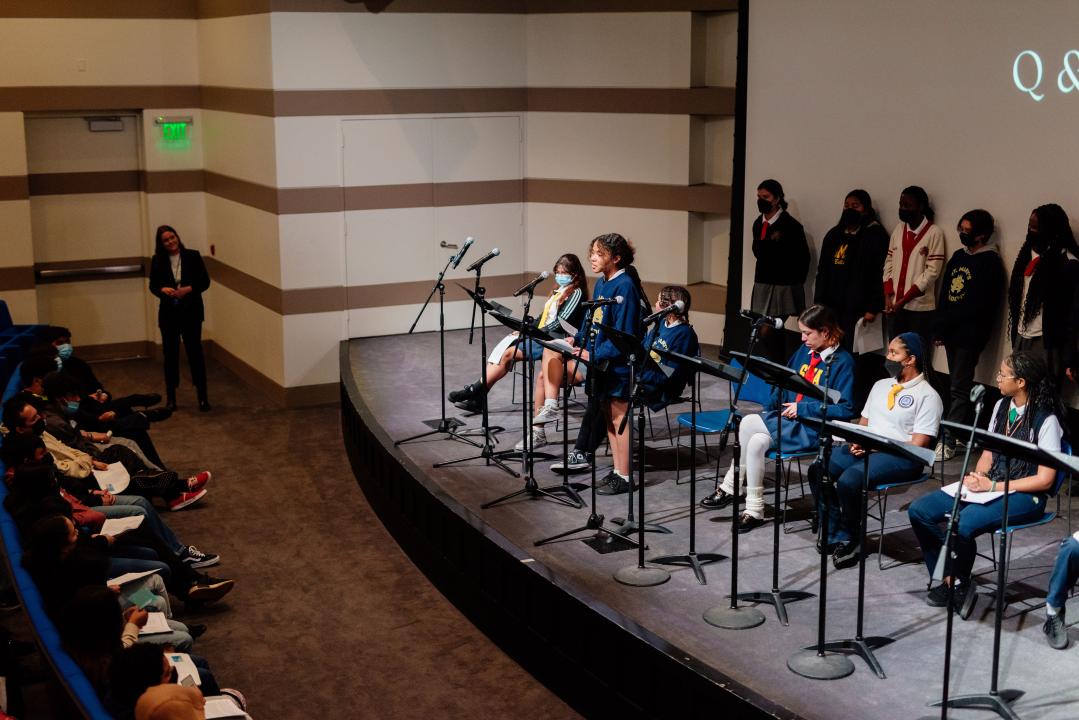 Photo of a group of students lined up on a stage. One student is standing in front of a microphone speaking to the audience.