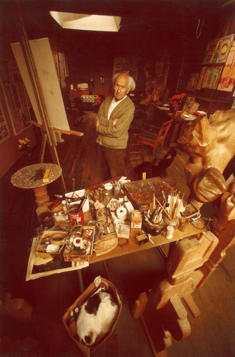 Photo of Peter Krasnow in his studio standing by an easel amongst materials and clutter