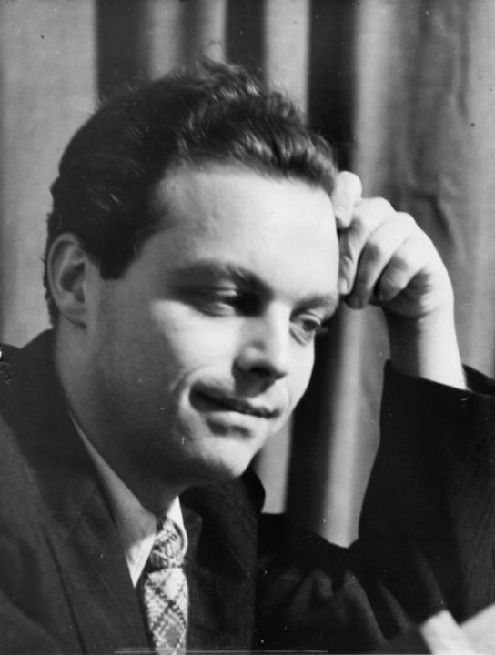 Black and white photo of composer Lou Harrison sitting in front of a curtain looking down, away from the camera