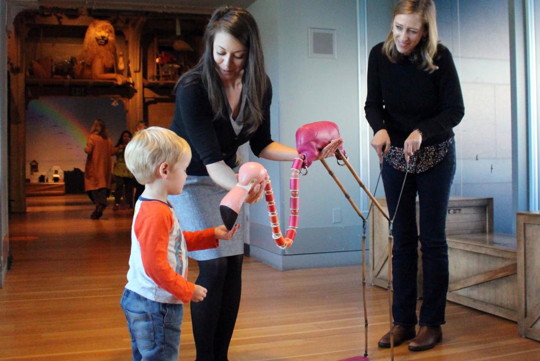 Two puppeteers engage a young child with a pink flamingo puppet with a craved wooden head and spools of thread for a neck.