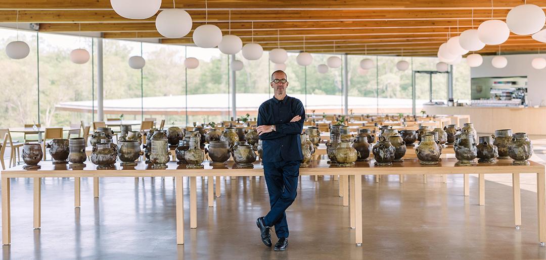 A man standing with arms crossed in front of large table in a large windowed room. The table has row and rows of pottery vessel on it.