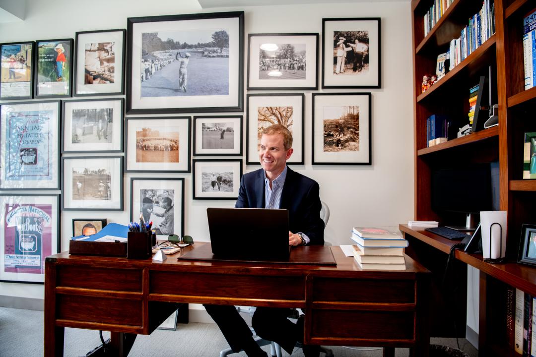 A smiling man sits behind a laptop computer at a wood desk, next to a wood book shelf. The wall behind him is adorned with photos.