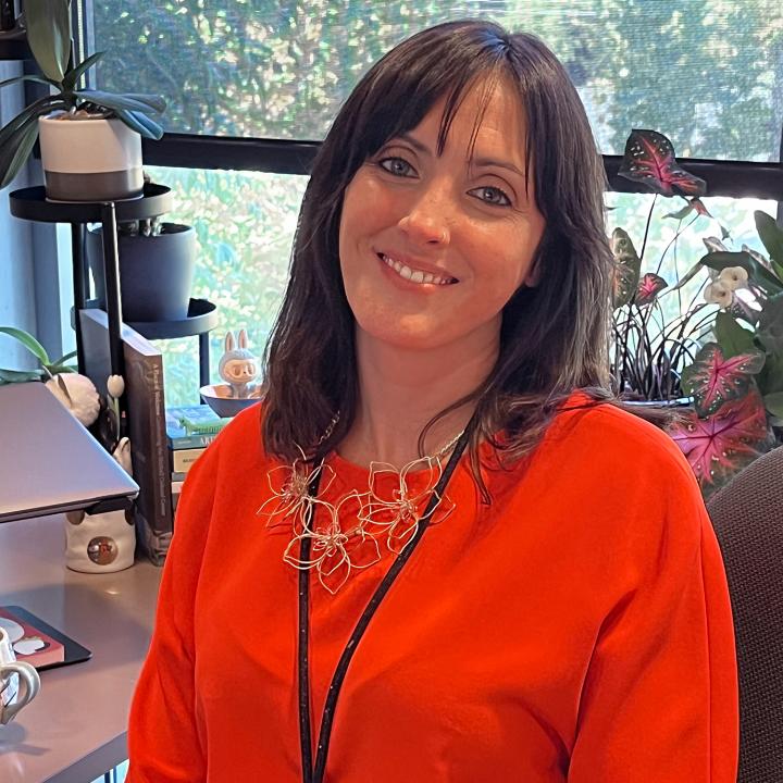A young woman with long brown hair smiles for a photo at her desk.