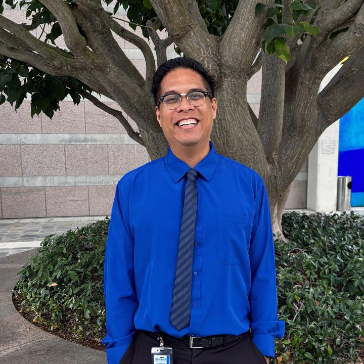 A mand wearing blue shirt and a tie, smiles for a photo under a tree.