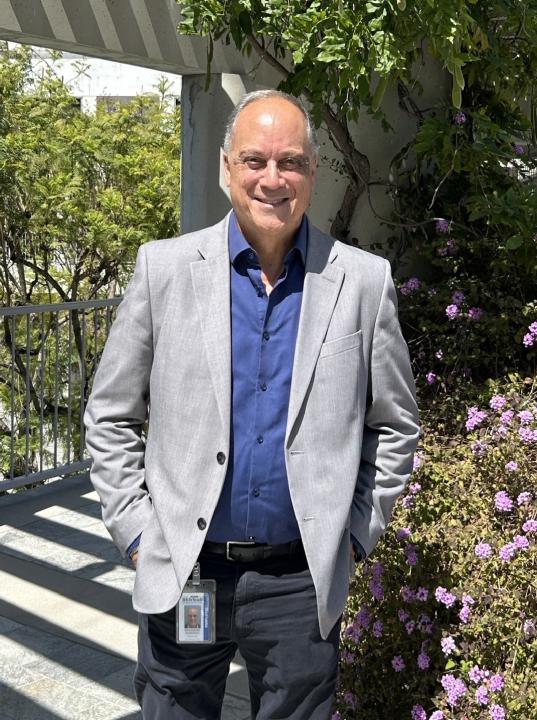 A man in a suit stands next to greenery.