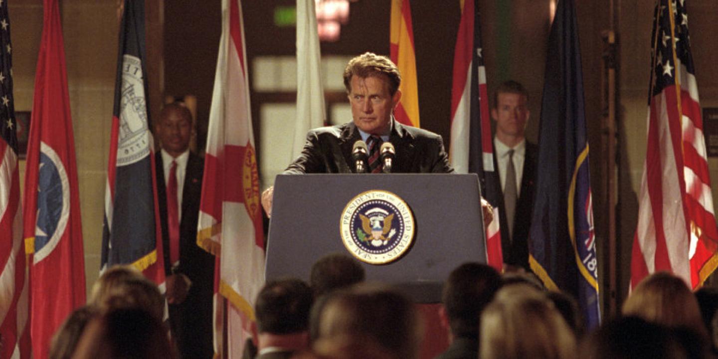 Image from the show The West Wing showing the president standing at a podium surrounded by flags and press