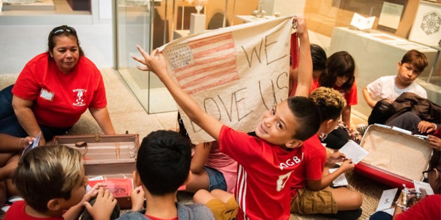 A teacher and students sitting on the floor, one boy holds up a banner with a US flag drawn on it and the words, We Love Us.