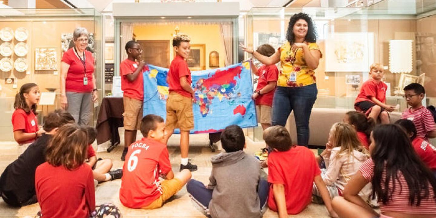 a teacher standing in front of students sitting on the floor; other students hold up a world map