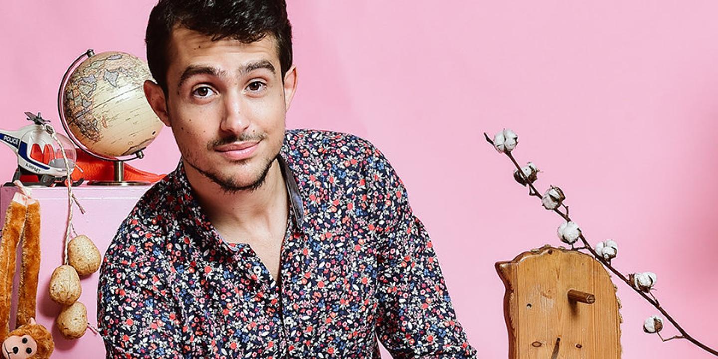 Headshot of Guy Mintus sitting in front of a pink background and surrounded by various, whimsical toys and items