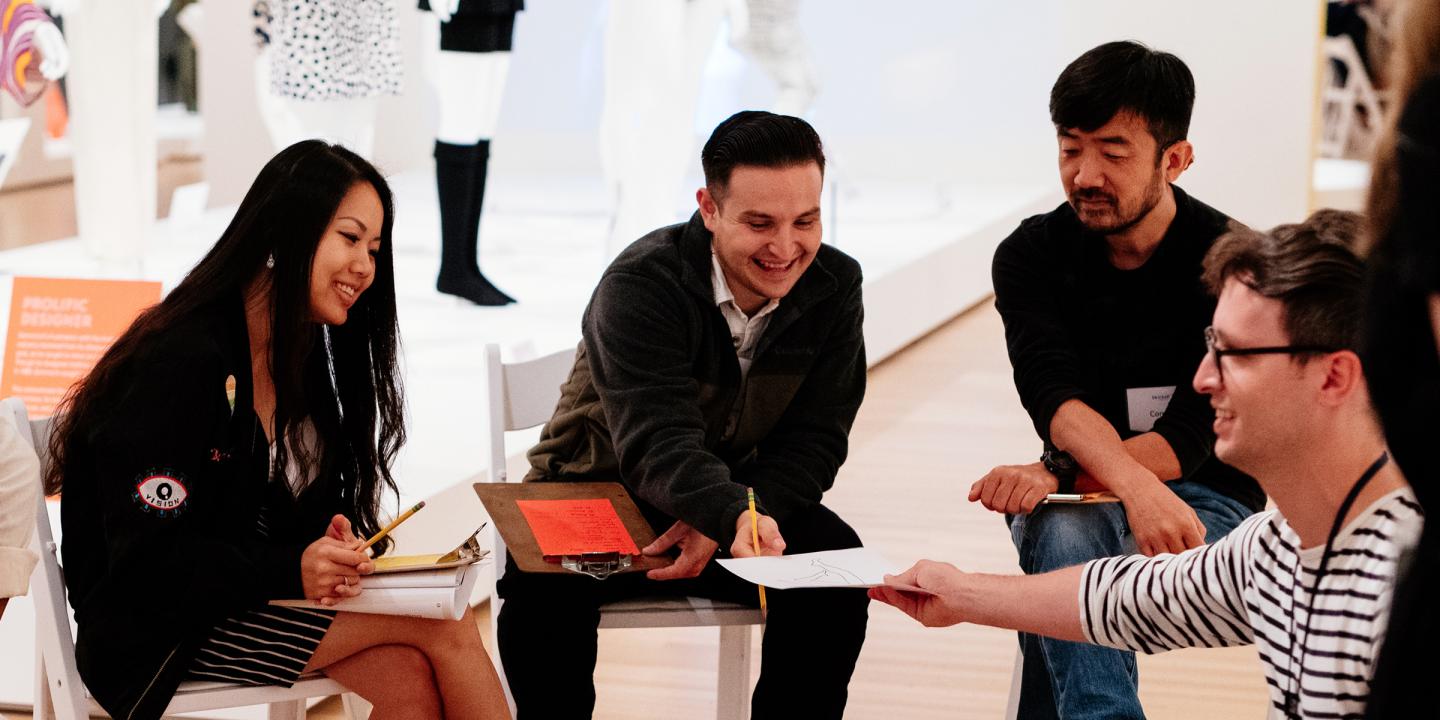 Four adults with clipboards and pencil in a circle looking at a drawing in a gallery.
