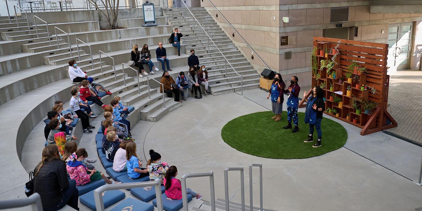 Three educator performing a story in front of an audience kids and adults in an outdoor theater. There are standing on green grass-like circular matt with a wooden backdrop with colorful potted plants.