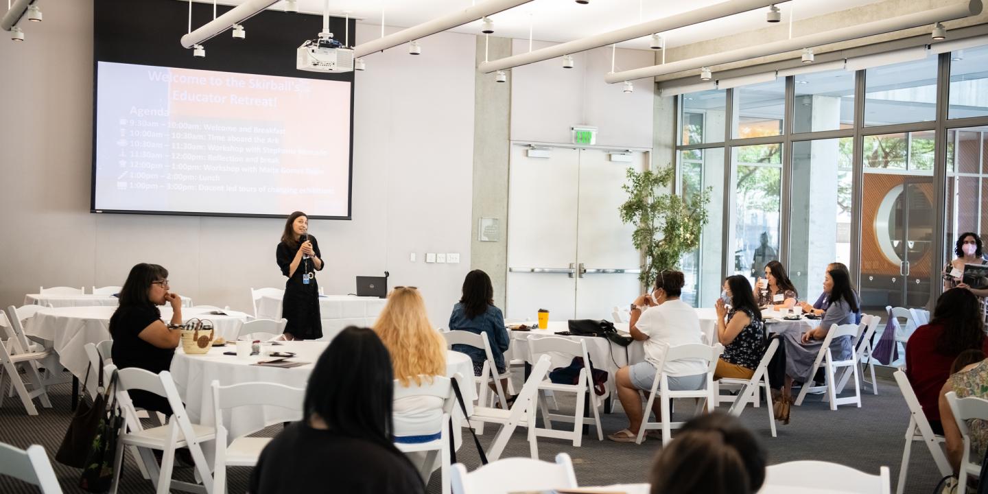 Photo of a large conference room. People are sitting at round tables watching a woman standing with a microphone. A screen with a slideshow is behind the standing woman.