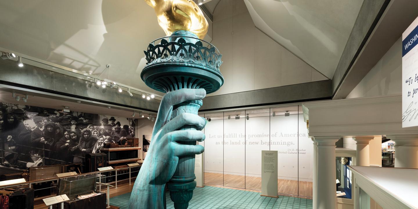 Photo inside the exhibition Vision and values showing a replica of the statues of liberty hand holding the torch among a large photo mural of immigrants on Ellis Island