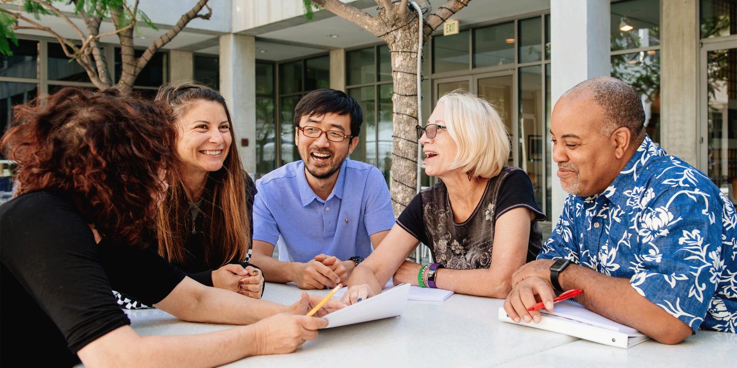 A photo of five people sitting together at a table outside. They are holding papers and pens while laughing with each other.