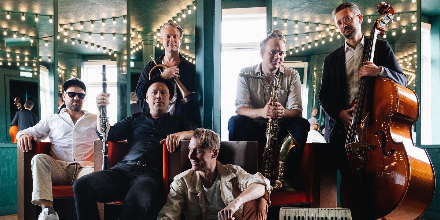 Photo of the six member band sitting in a mirrored room. Each member is holding their instrument and smiling at the camera.