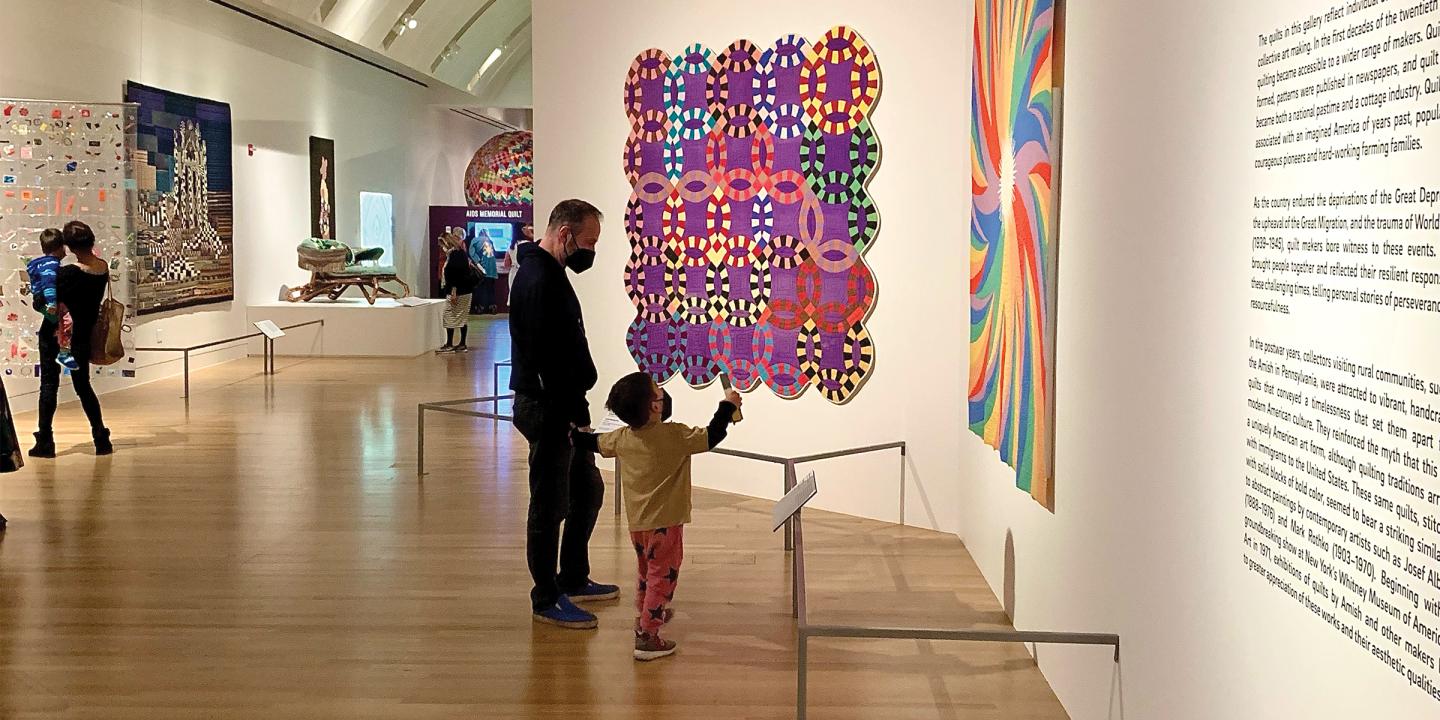 Photo of a museum gallery. Large, colorful quilts are hung on white walls and a myriad of people are looking at them. In the center of the photograph are a father and young son looking at a quilt together.