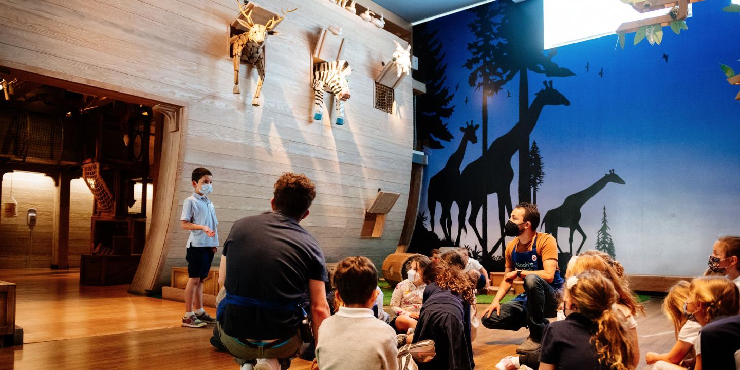 Kids gathered, sitting on the floor with attention on one child speaking. In the noah's Ark rainbow gallery with a wooden ark ing the background and animals hanging of the ark.
