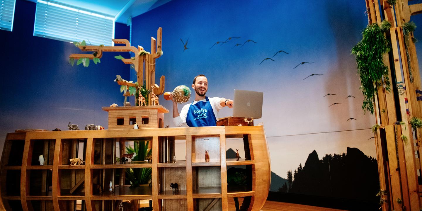 a Noah's Ark educator shaking a musical gourd telling a story to a computer screen from the Naoh's Ark gallery with a sky wallpaper and wooden ark.