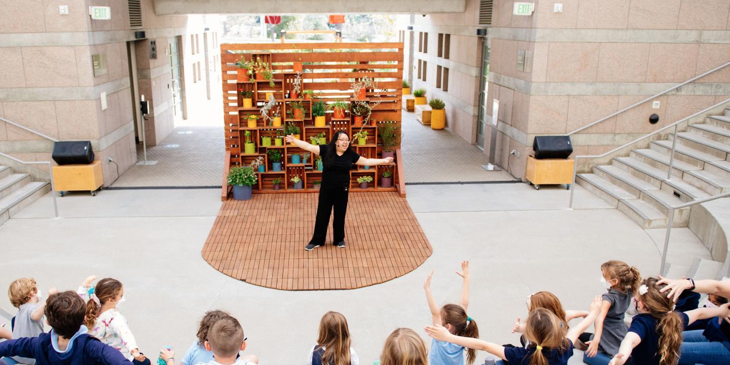 Educator in performing a story with arms stretched wide while children are watching from outdoor amphitheater seats. There is a wooden planked backdrop with color potted plants.