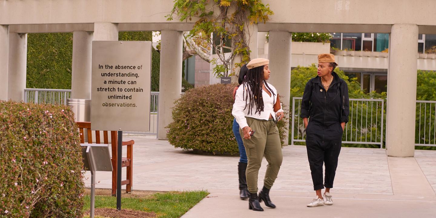 Photo of a group of people walking outside on the Skirball campus. In the foreground an artwork with the words, "In the absence of understanding, a minute can stretch to contain unlimited observations."